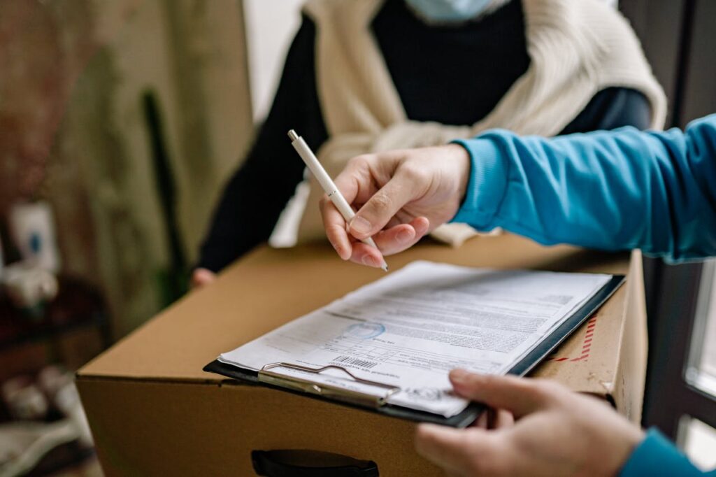 pexels photo 6818154 Close-up of a person signing a delivery document placed on a large cardboard box indoors.